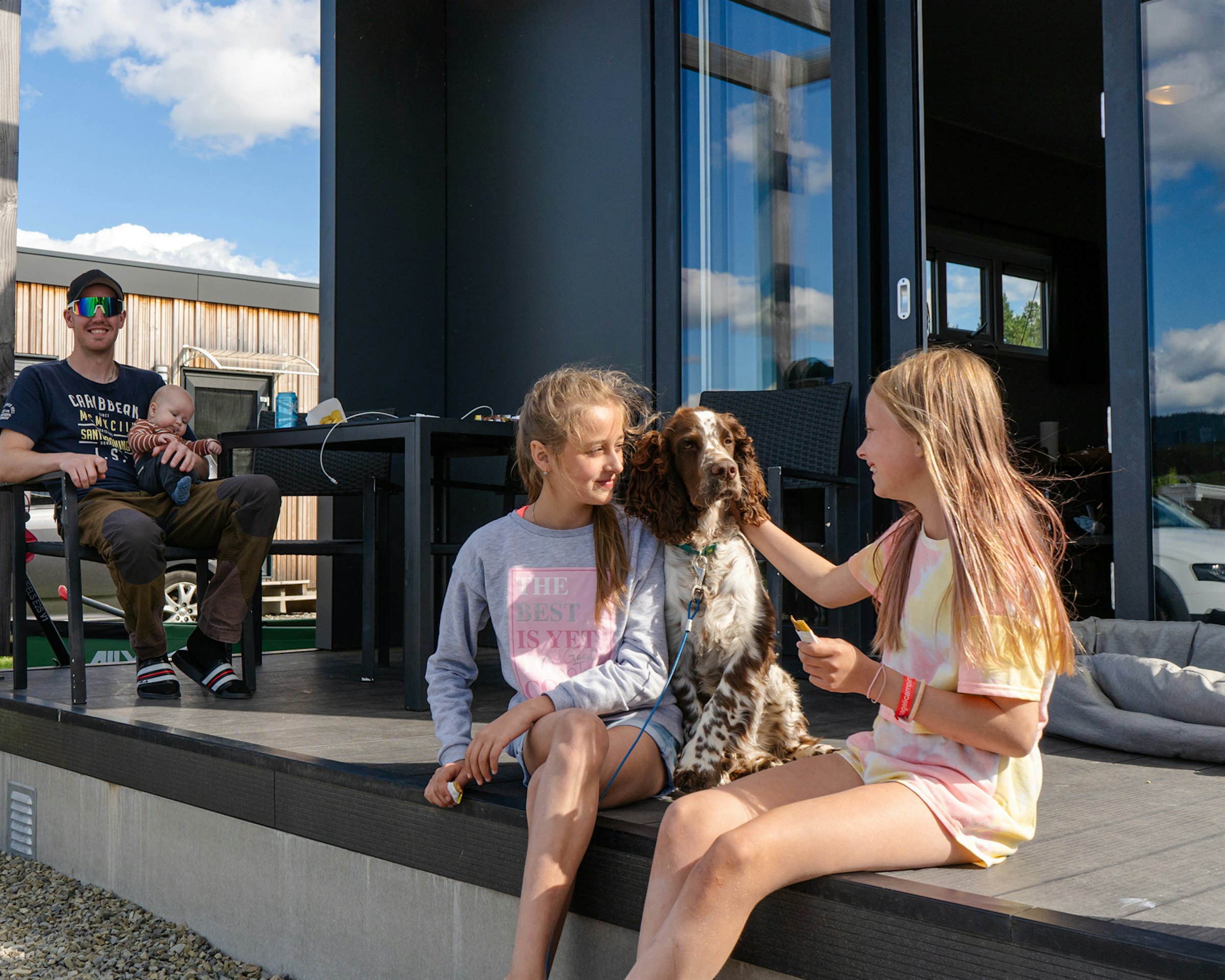 Zwei Mädchen sitzen lächelnd auf der Veranda und streicheln einen Hund, Papa sitzt im Hintergrund mit einem Baby auf dem Schoß und lächelt. Foto