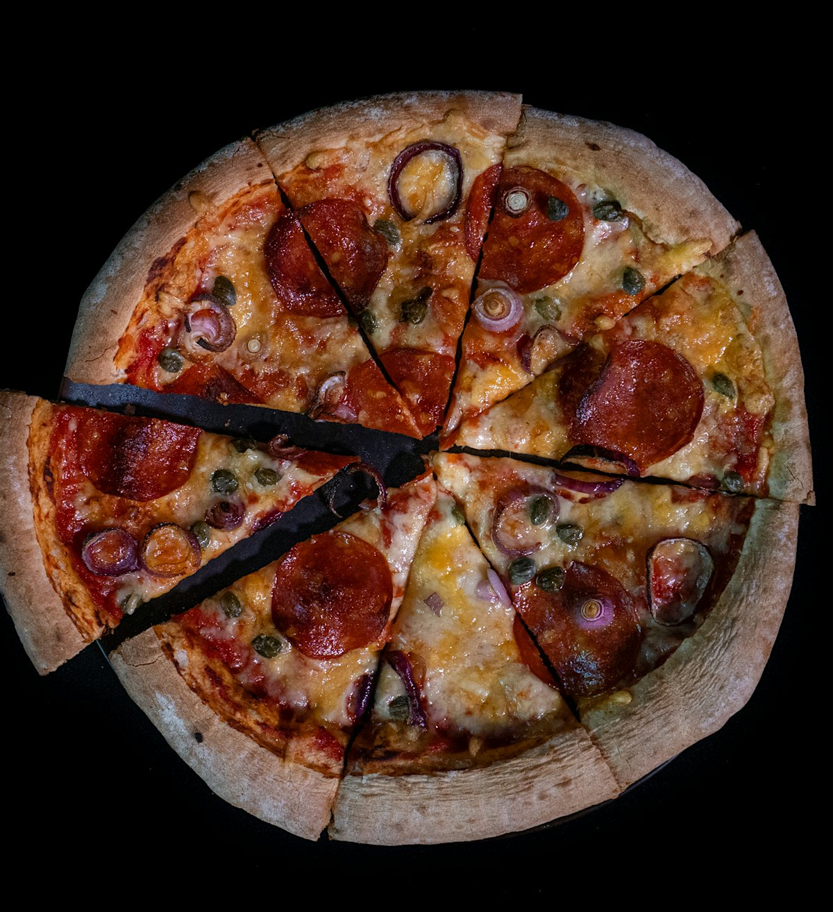 Pizza sett ovenfra på svart bakgrunn. Pizzaen har en tykk kant, ost, grønnsaker og pølsebiter på toppen, Den er delt i åtte trekanter hvor den til venstre er tatt litt ut fra ringen.