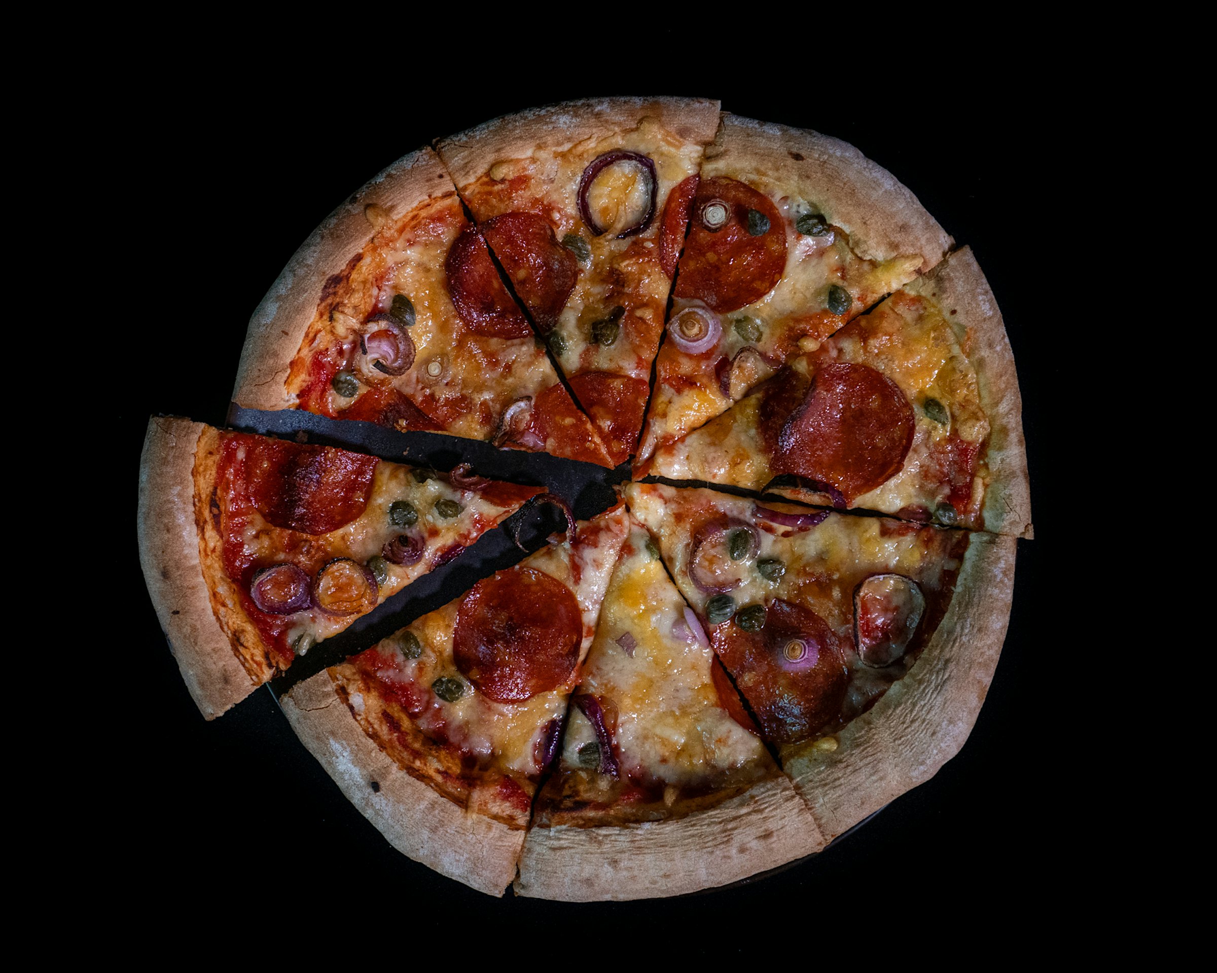 Pizza sett ovenfra på svart bakgrunn. Pizzaen har en tykk kant, ost, grønnsaker og pølsebiter på toppen, Den er delt i åtte trekanter hvor den til venstre er tatt litt ut fra ringen.