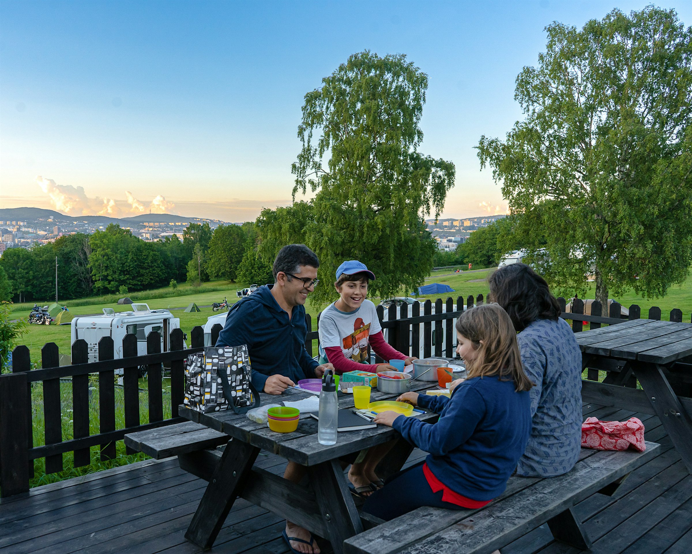 Familie spiser middag ute på en terrasse på benker med bord i kveldssola. Mann og gutt sitter vendt mot kvinne og jente som sitter med ryggen vendt mot kamera. Bak ser man gresslette, skog og Osloby.