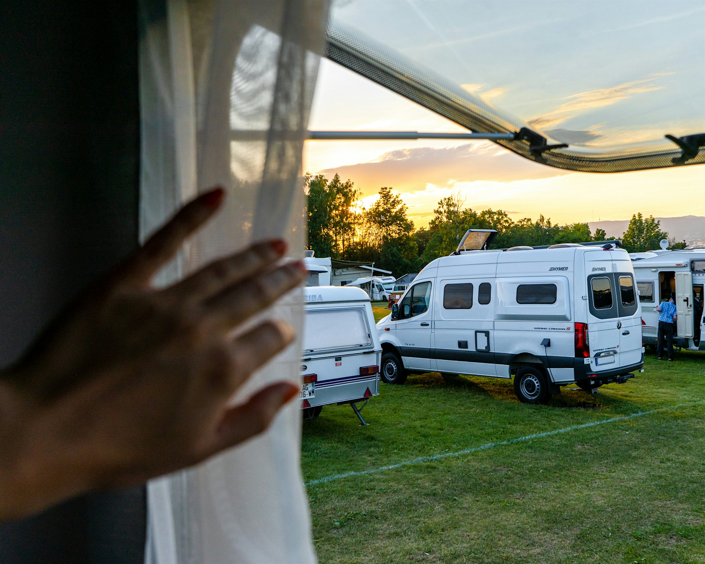 Hånd skyver tynn gardin til siden i campingvinduet. Man ser utover campingbiler på gresslette i solnedgang.