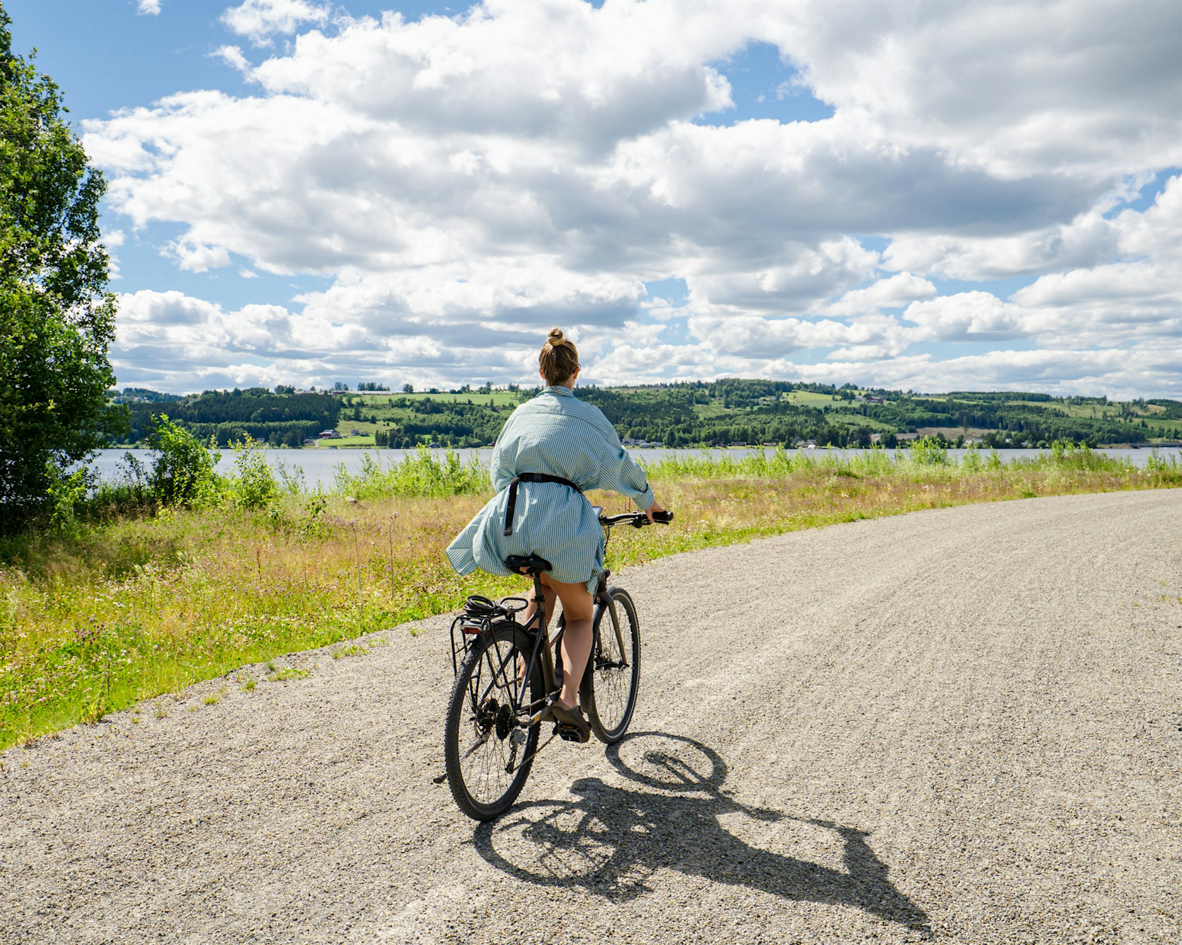 Girl cycling on footpath surrounded by grass, and Mjøsa with forest in the background. Photo