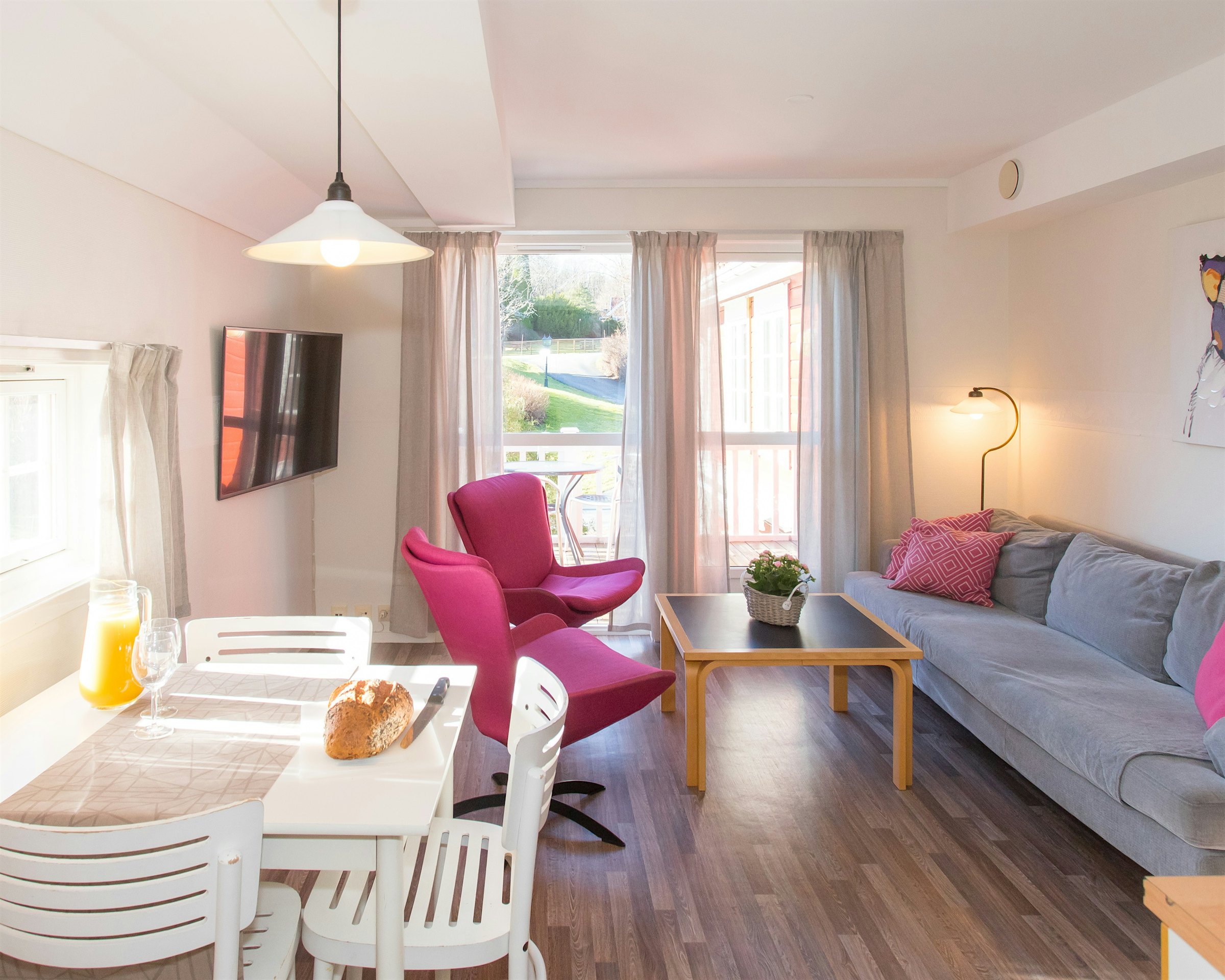 Heller und offener Raum mit viel natürlichem Licht durch große Fenster. Zwei rosafarbene Sessel, Sofa, Fernseher und gedeckter Esstisch zum Frühstück. Foto