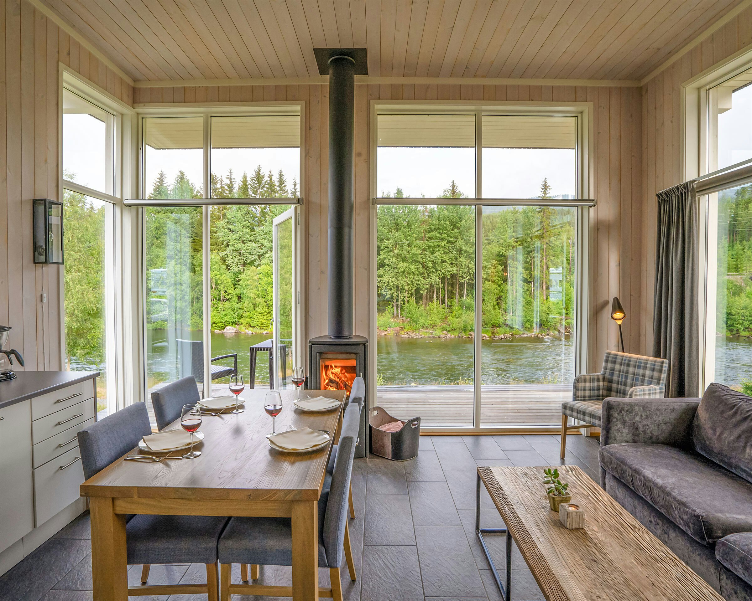 Lys stue med vinduer fra gulv til tak, spisebord sofa og peis. Utsikt over grønne trær og elv.