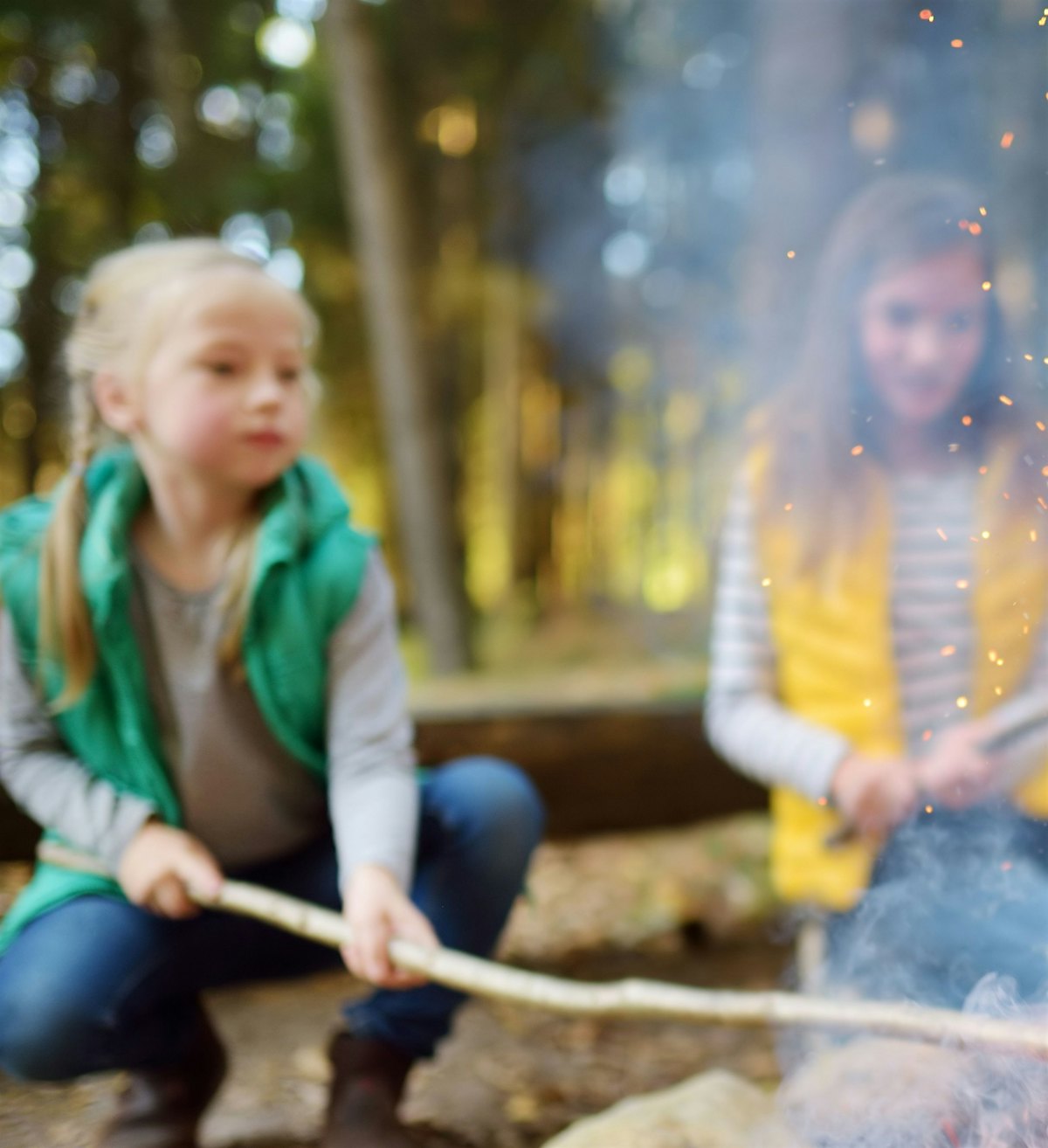 I skogkanten sitter to uklare jenter med pinner over et bål. Det er flammer og gnister i forgrunnen.