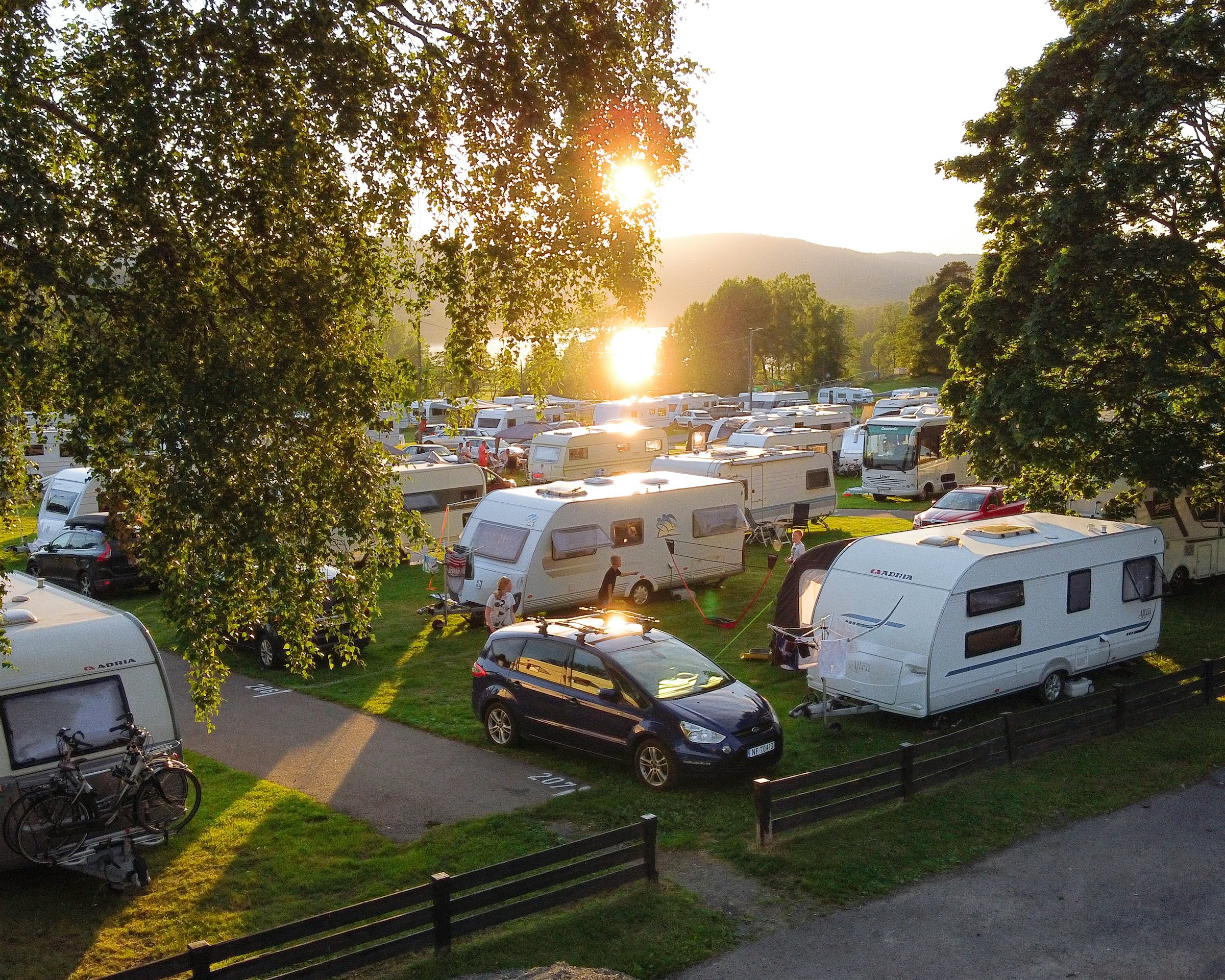 Mange campingvogner står oppstilt på campingplass, med solen som går ned i bakgrunnen. Foto