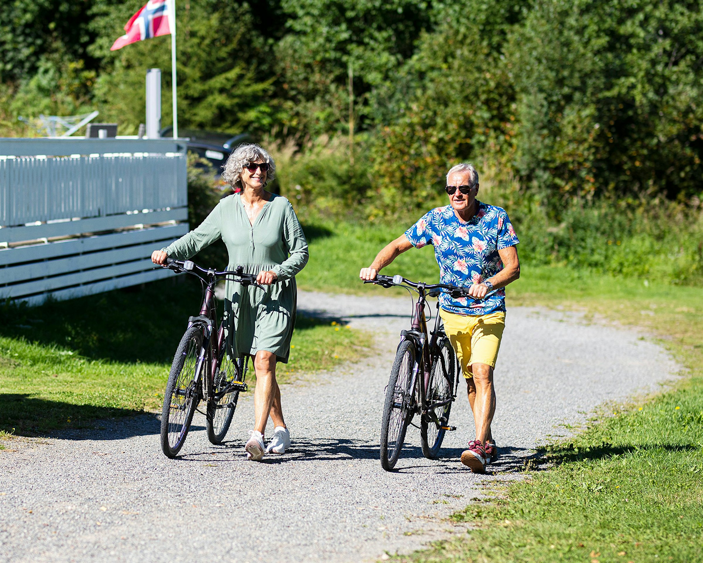 Et eldre par sykler på grusvei ved Topcamp Mjøsa i sommersolen. Foto