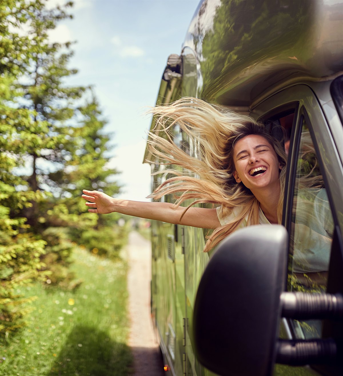 Blonde Frau am Fenster eines Wohnmobils mit ausgestreckten Händen lächelnd und genießt die Fahrt.
