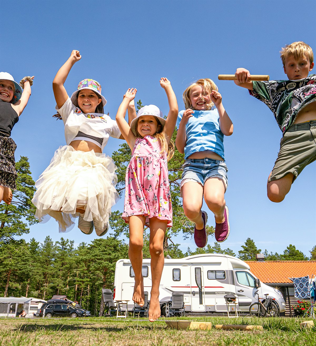 A group of children jump in the air with their hands above their heads at a campsite. Everyone smiles big. Photo