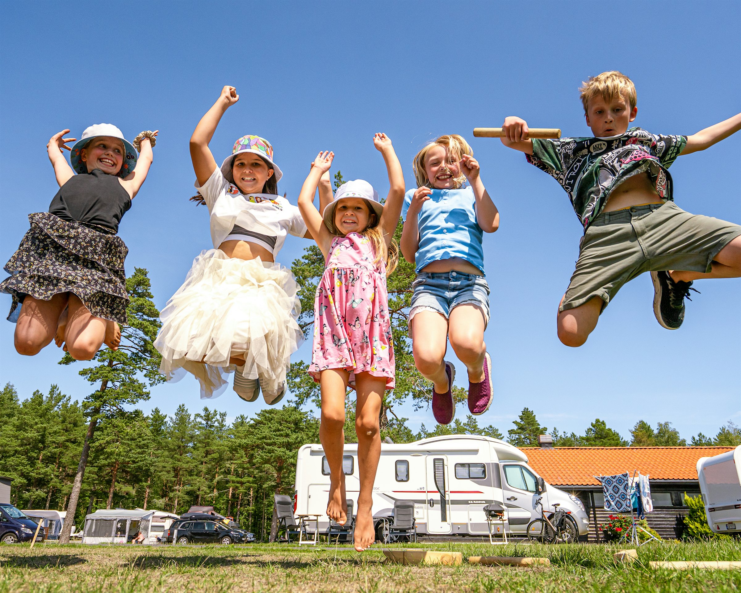 A group of children jump in the air with their hands above their heads at a campsite. Everyone smiles big. Photo