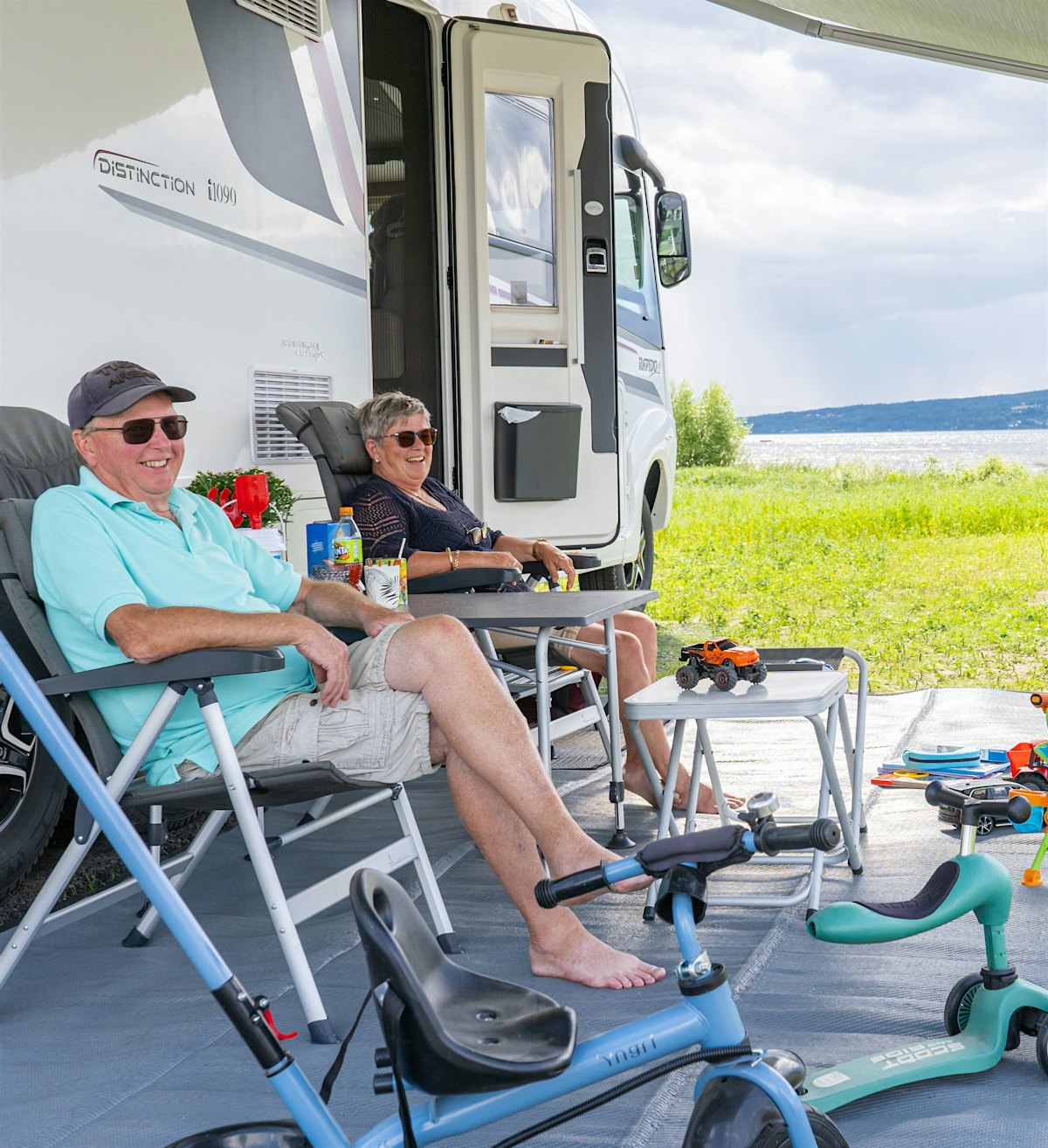 A grandfather, a grandmother and a boy are sitting outside a mobile home and enjoying themselves. Two sitting scooters are in the foreground, water and grass in the background. Photo