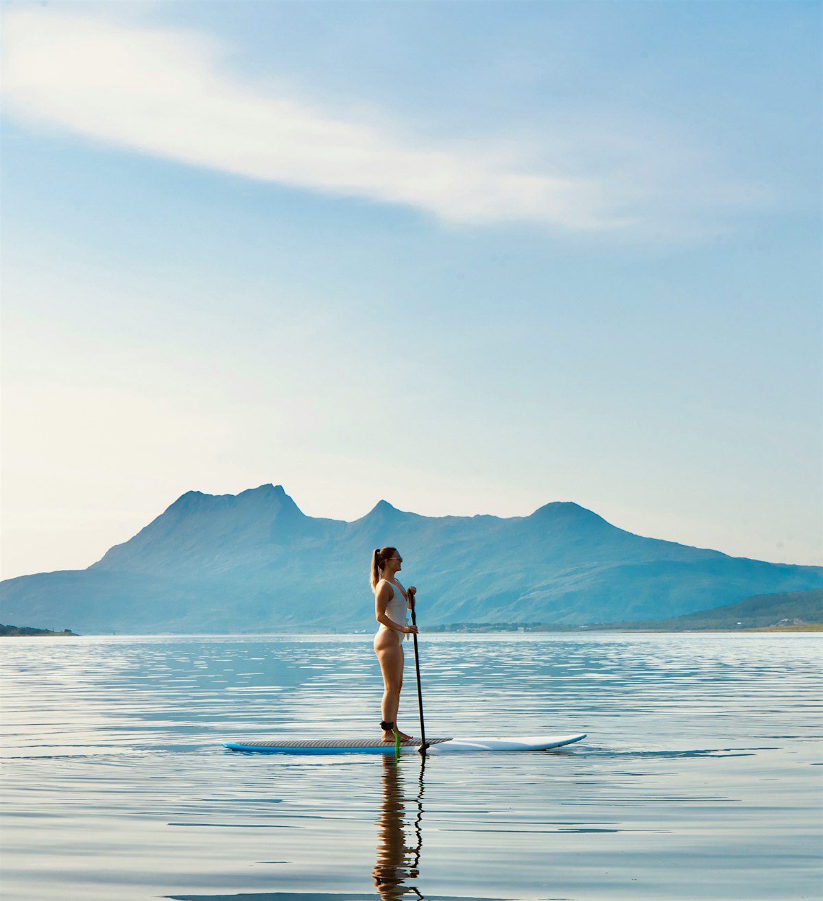 Girl stands upright on a SUP board with mountains in the background. Photo