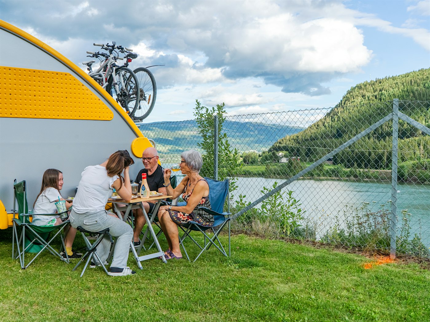 Family eating dinner outside caravan, with river and mountains in the background. Photo