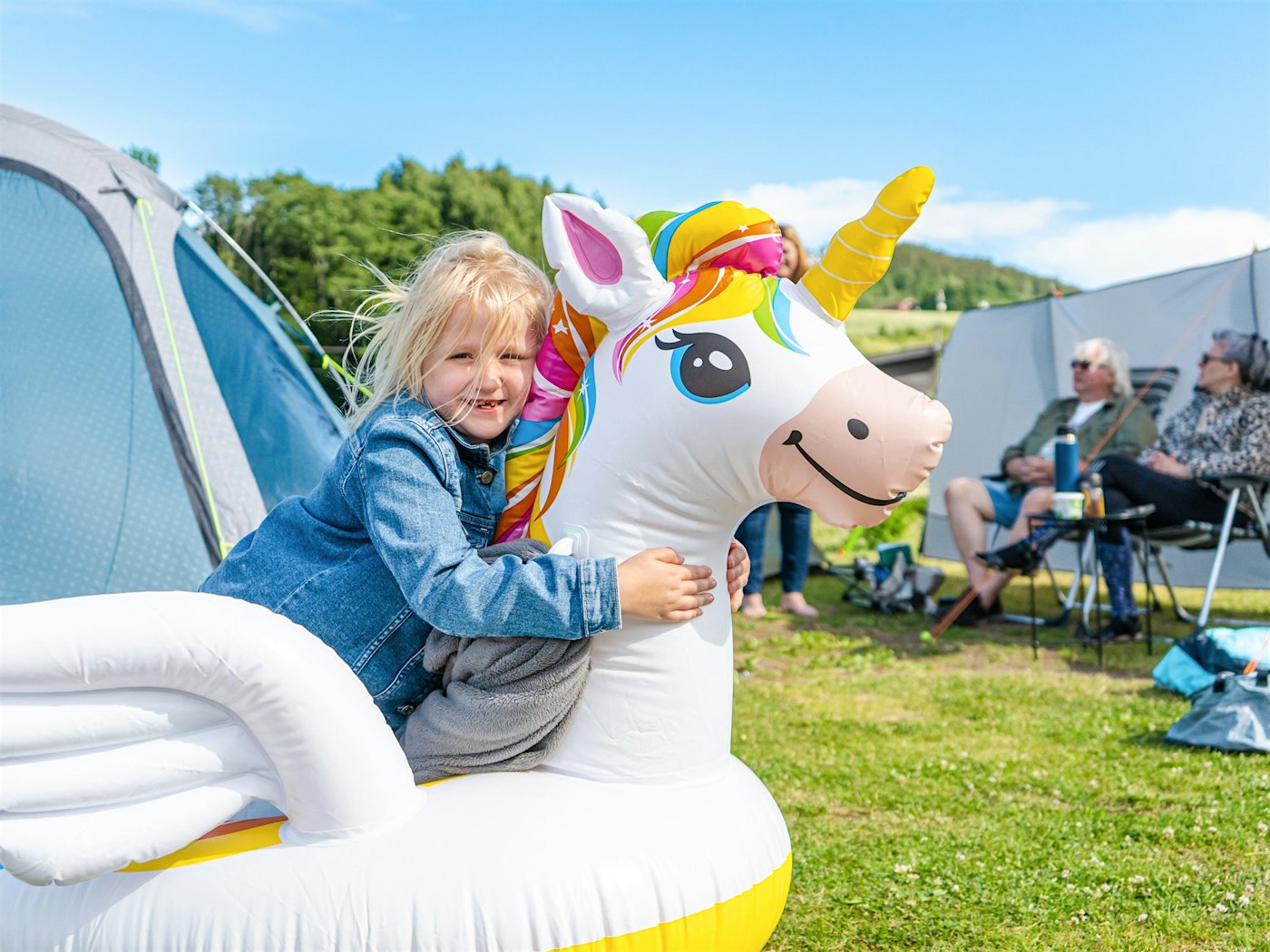 Girl sits on a unicorn bath toy and smiles. Caravan and tent in the background. Photo