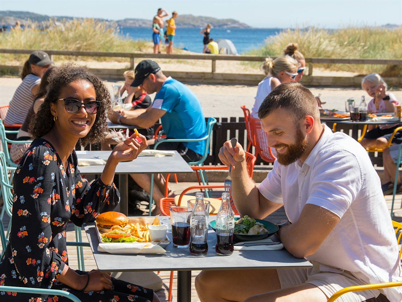 Mann og kvinne sitter og spiser på uteservering. I bakgrunnen ser man stranden. Kvinnen smiler mot kamerat, mannen ler ned mot bordet.