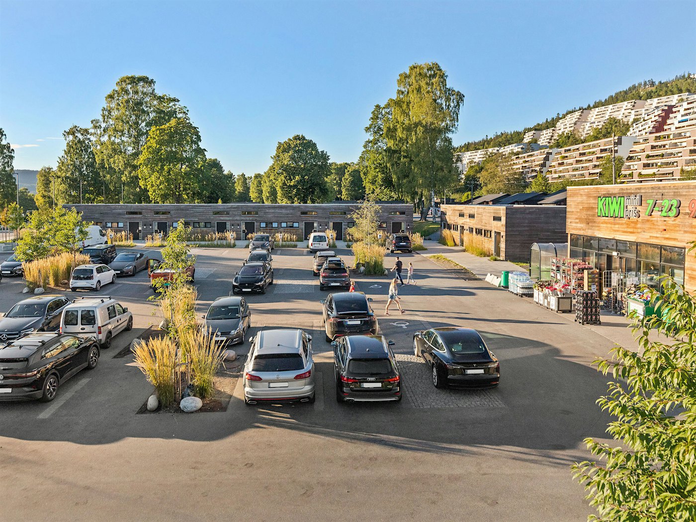Parkeringsplass med flere biler, leilighetsbygg i tre i bakgrunnen. Kiwi-butikk på høyre side. Det er trær på parkeringsplassen og i bakgrunnen.o
