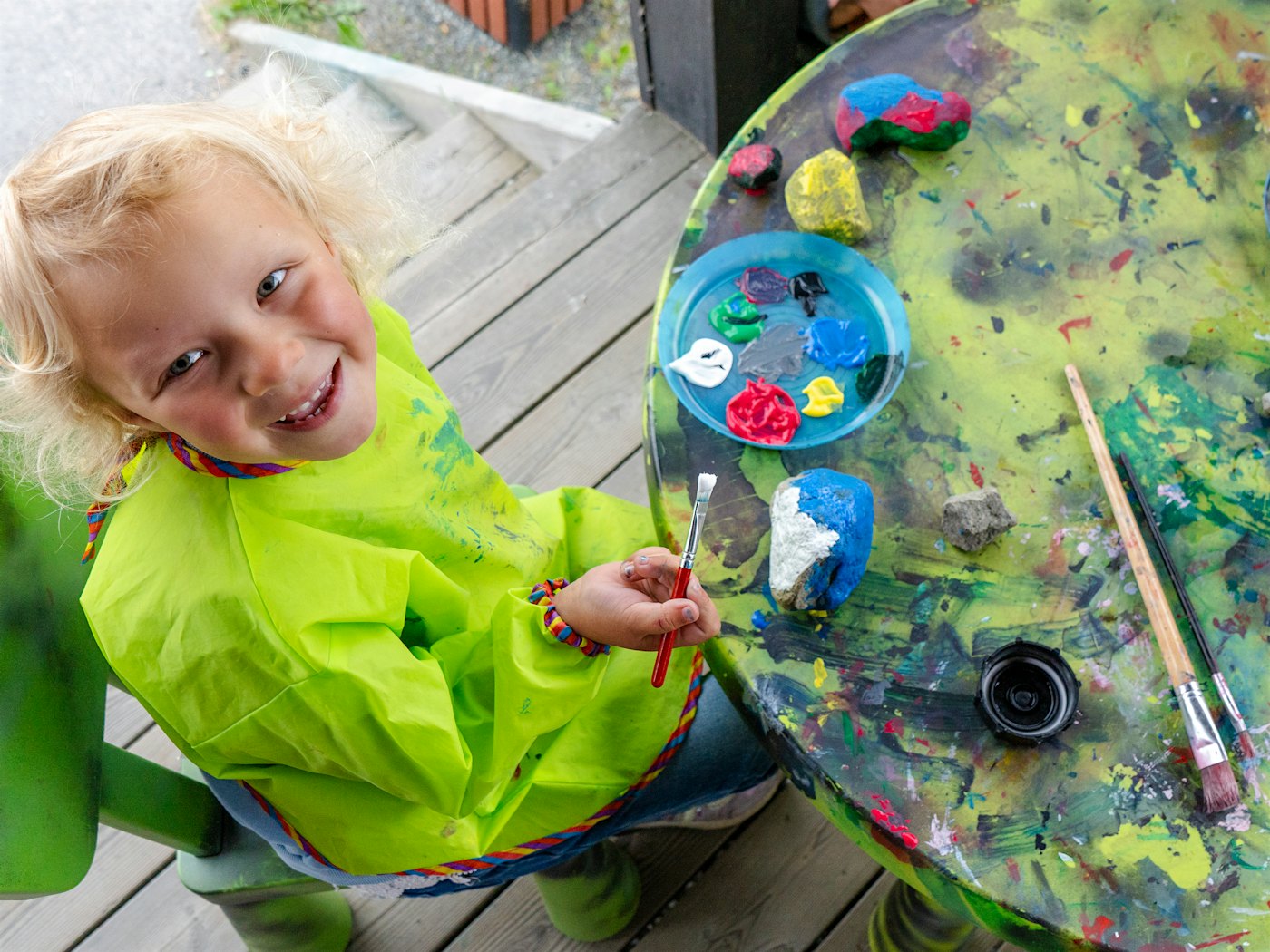 A child with a painting bib smiles and paints on a stone. Photo