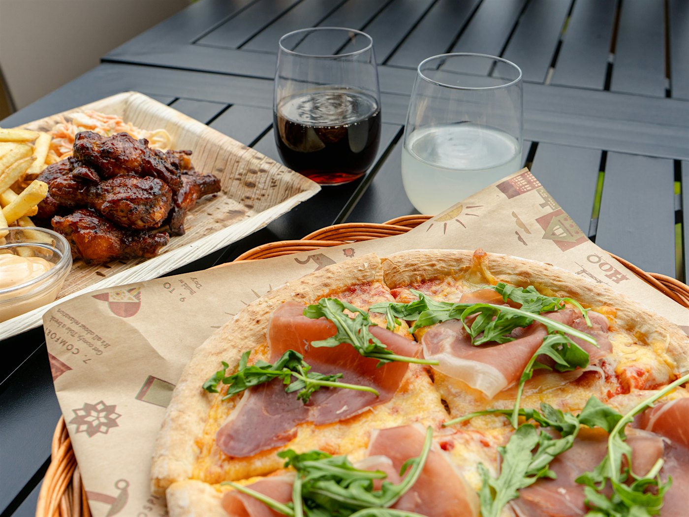 Pizza på pizzbrett med parmaskinke og ruccola halvvei inn i bildet. Buffalo wings med pommes frittes, er halvveis inn i bildet til venstre. To glass med drikke. Alt på et svart bord.