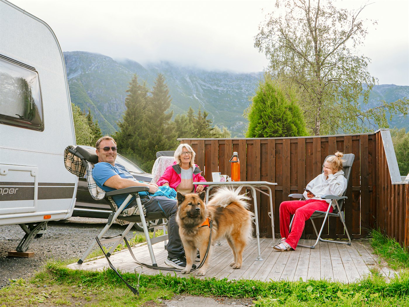 Mann, kvinne og barn sitter i hver sin campingstol rundt et bord vedsiden av campingvogn. Foran er det lys hund i bånd. Bak dem er det levegg, skog og fjell.