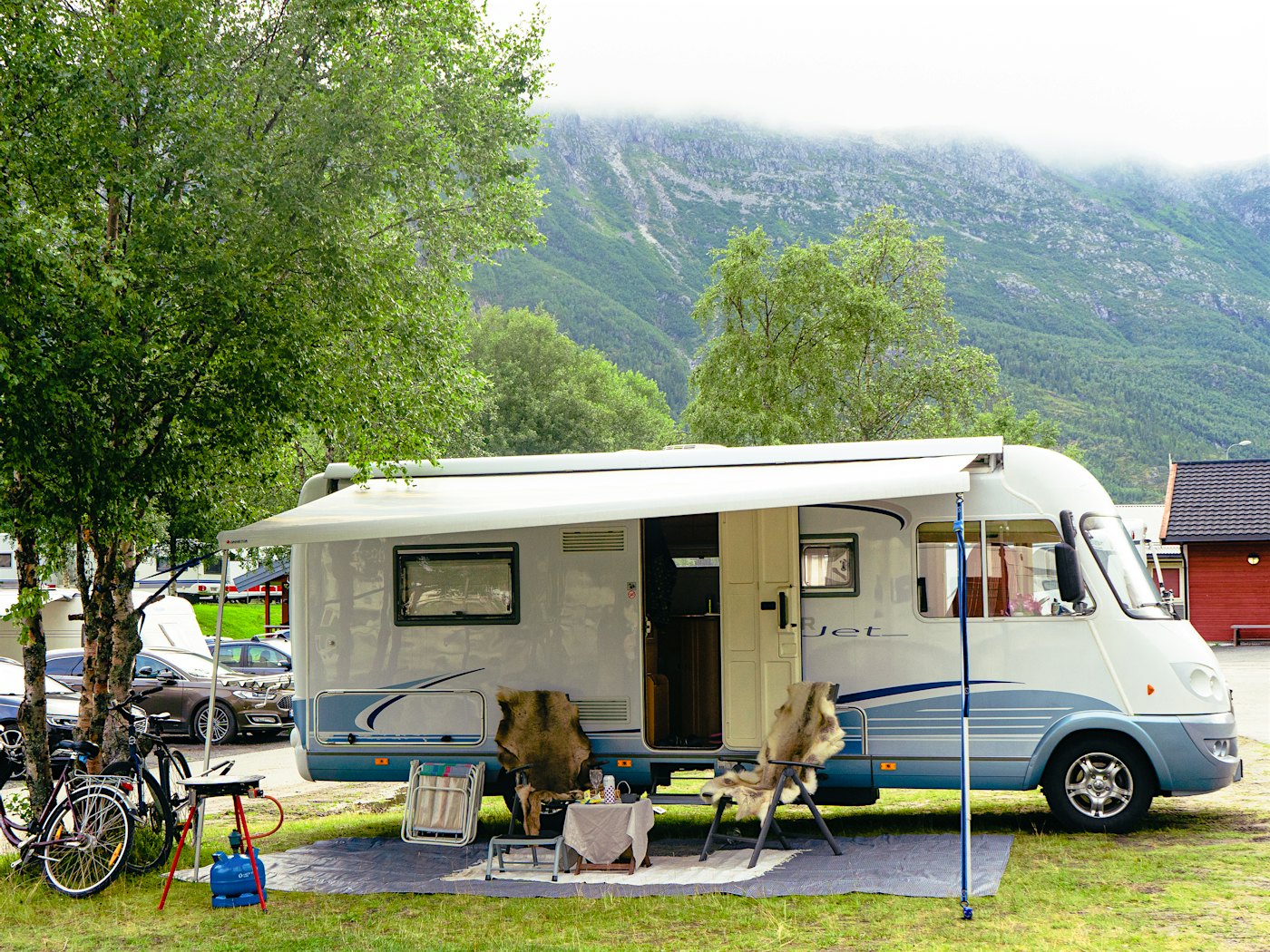 Motorhome with outdoor furniture and bicycles in front. Nicely located by a tree, with mountains in the background. Photo
