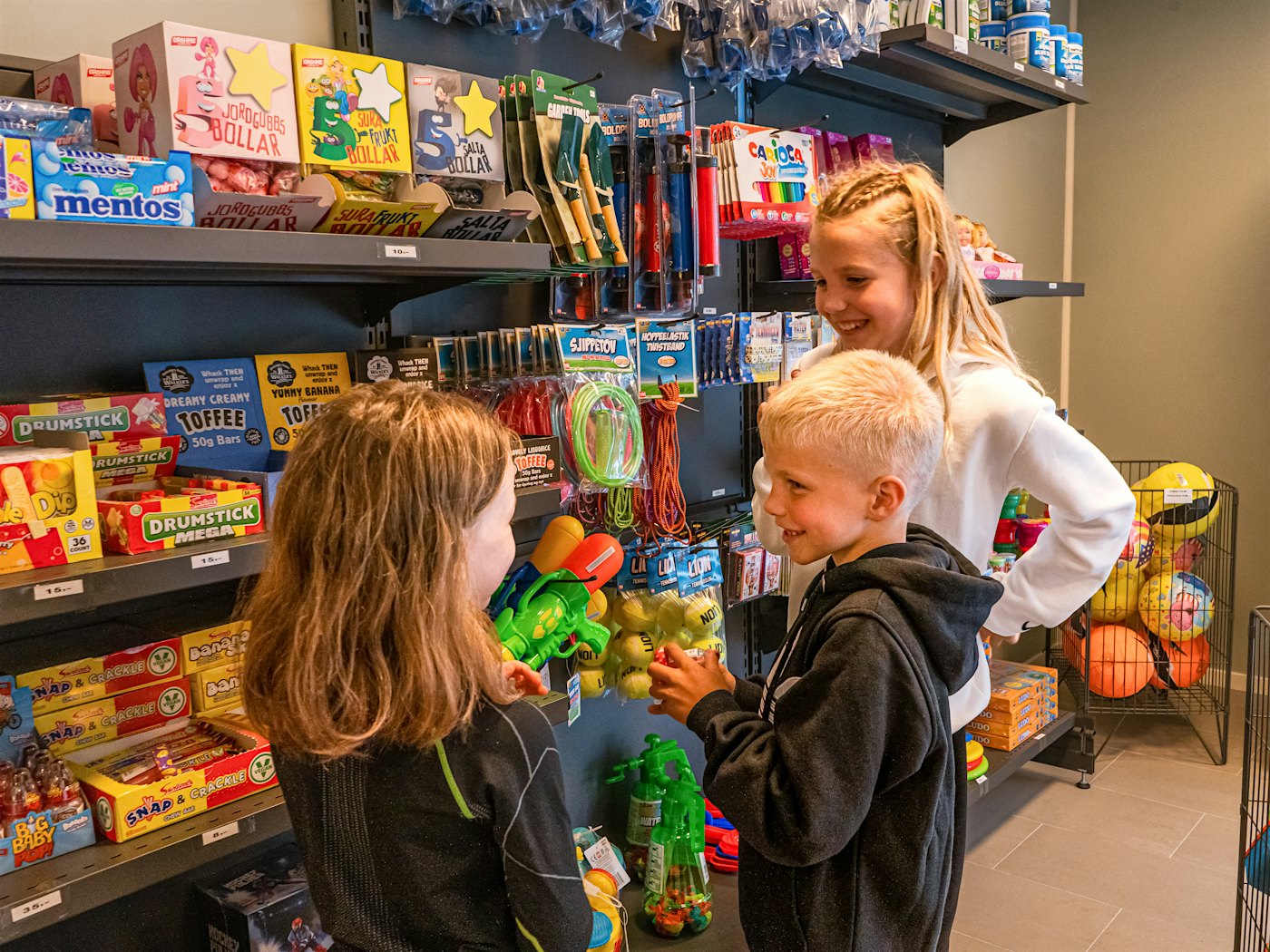 Tre barn står i butikk og ser på leker og godteri i hyllene.
