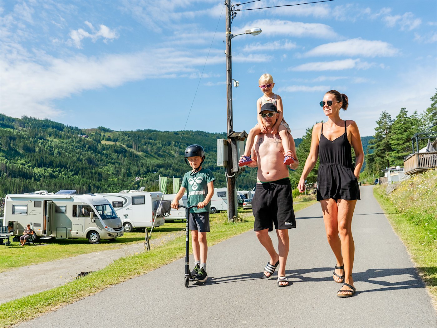 Familie schlendert lächelnd über den Campingplatz, Mädchen sitzt auf den Schultern des Vaters und Junge steht auf Roller. Foto