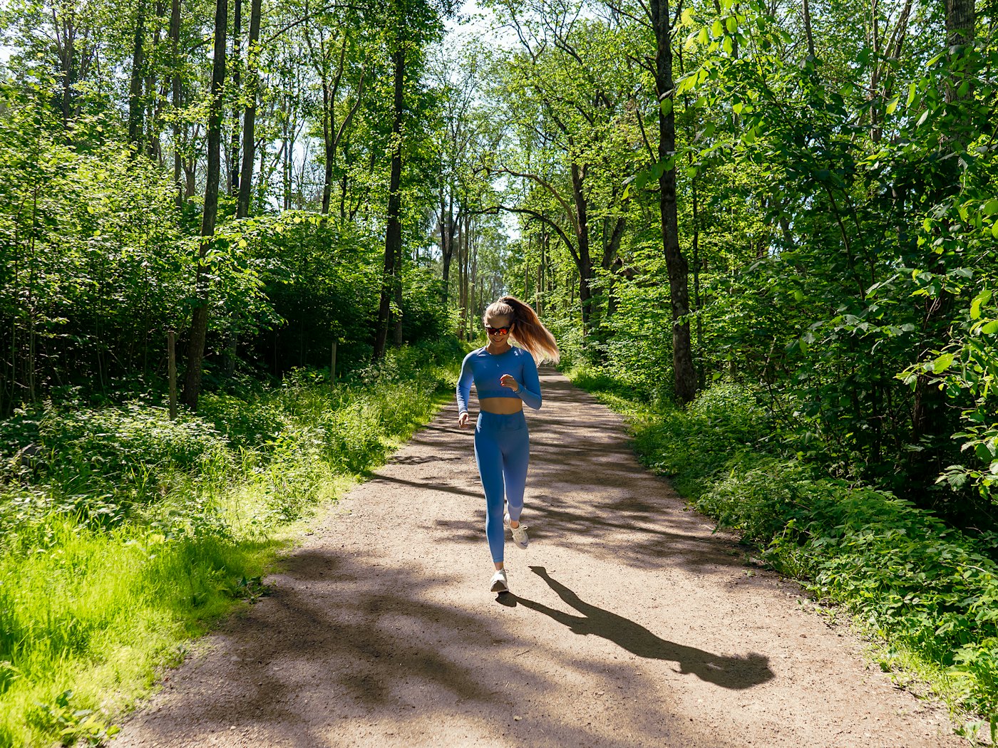 Dame i blått  jogger på en bred gruslagt sti i skogen mot kamera.