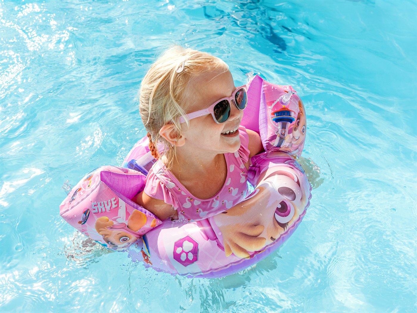 Jente med rosa solbriller flyter i badering og har på armringer i basseng.