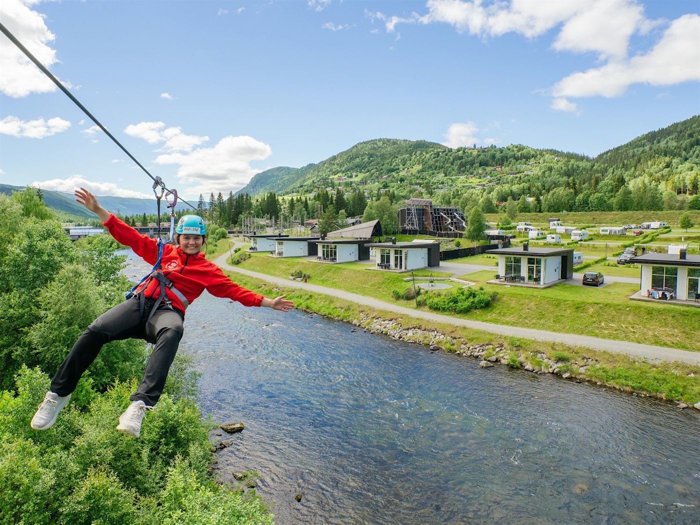 Lady in a zipline over the river, with the cabins and campsite in the background. Photo