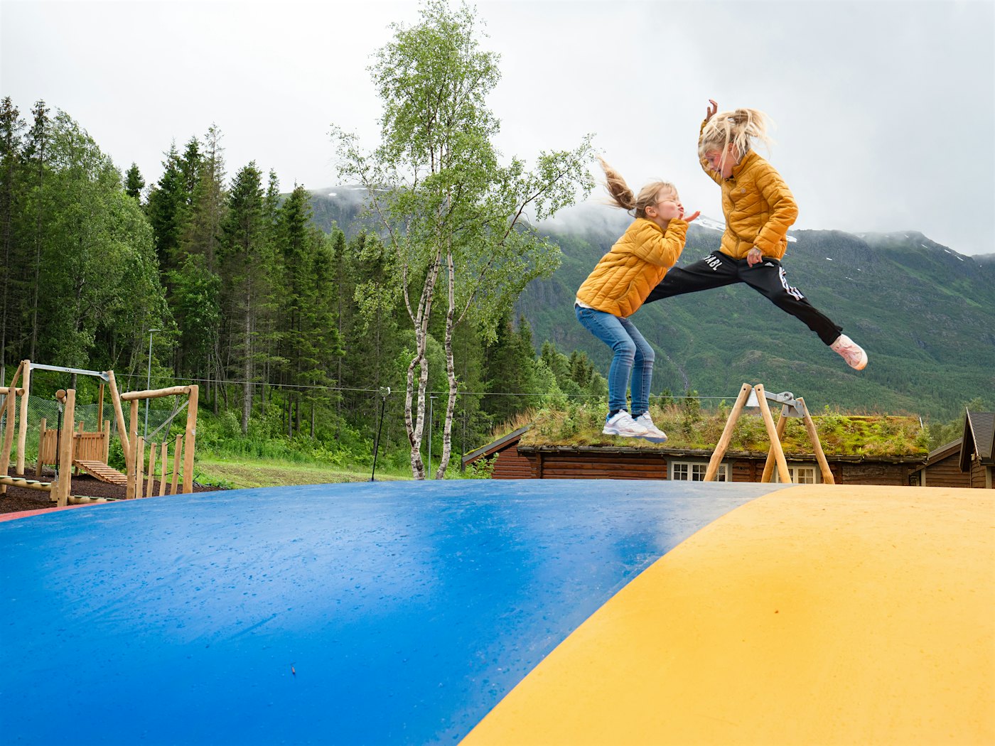 To jenter hopper på hoppepute med lekeplass og fjell i bakgrunnen.