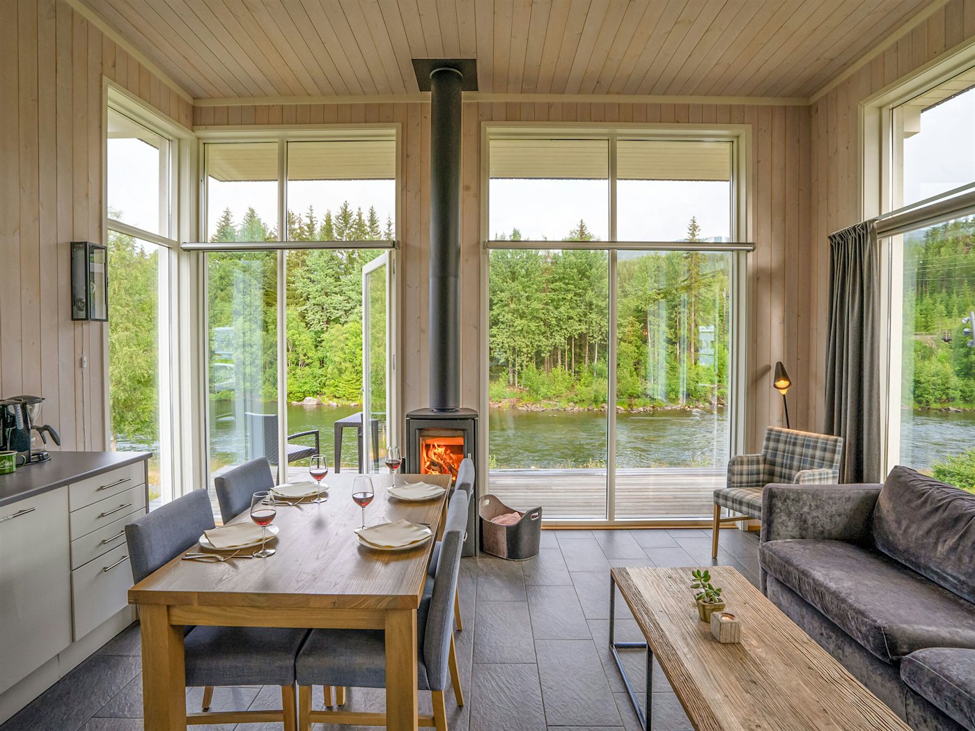 Lys stue med vinduer fra gulv til tak, spisebord sofa og peis. Utsikt over grønne trær og elv. Foto