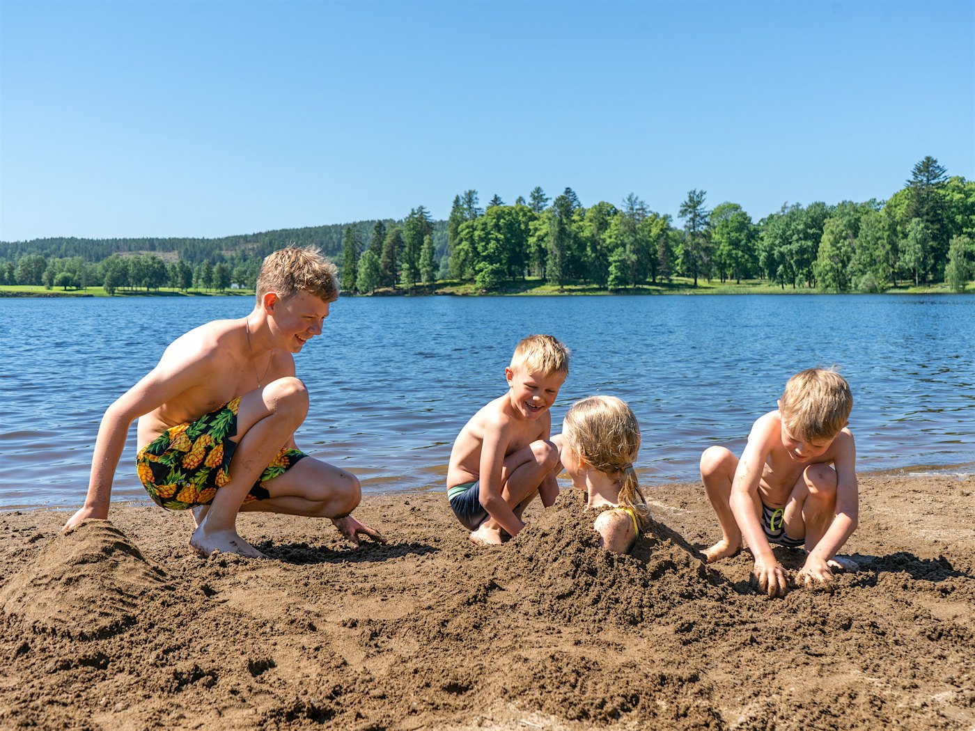 Far og to gutter graver ned jente i stranda. Hodet til jenten stikker opp. I bakgrunnen ser man blått vann og skog.