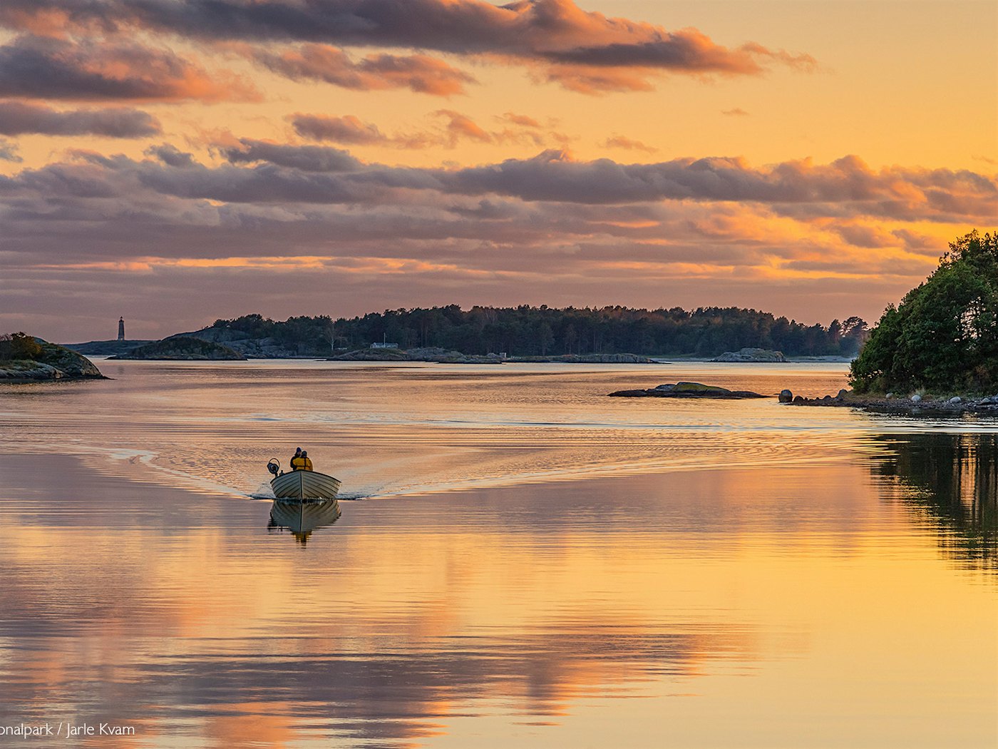 Båt kjører bortover blikkstille vann med dramatisk solnedgang som reflekteres i vannet.