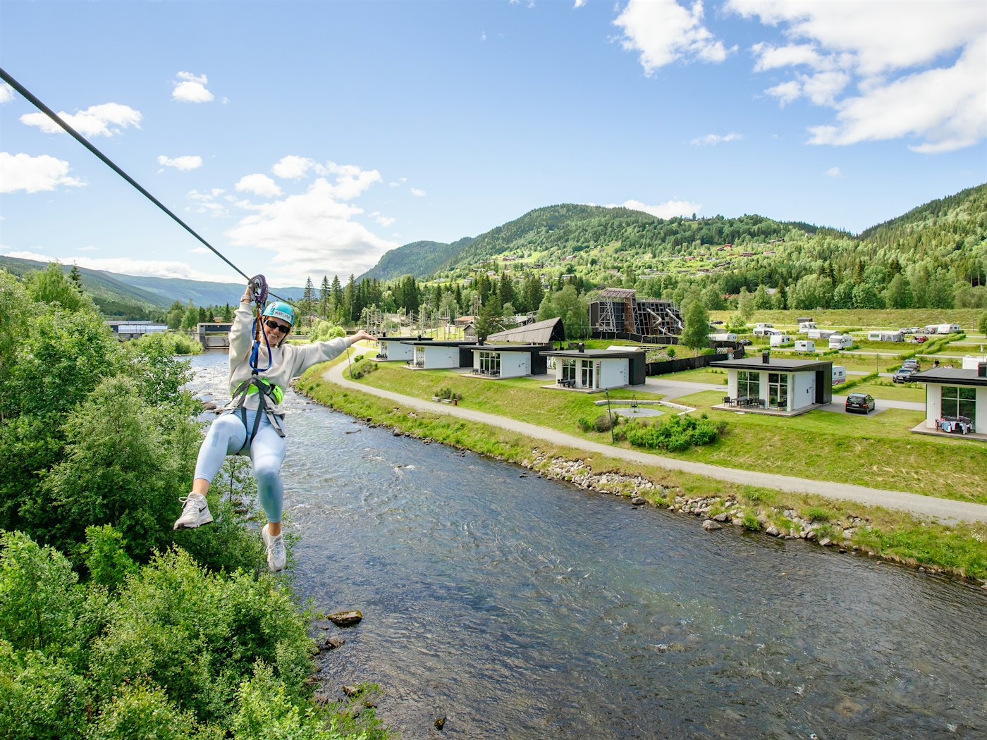 Lady in a zipline over the river, with the cabins and campsite in the background. Photo