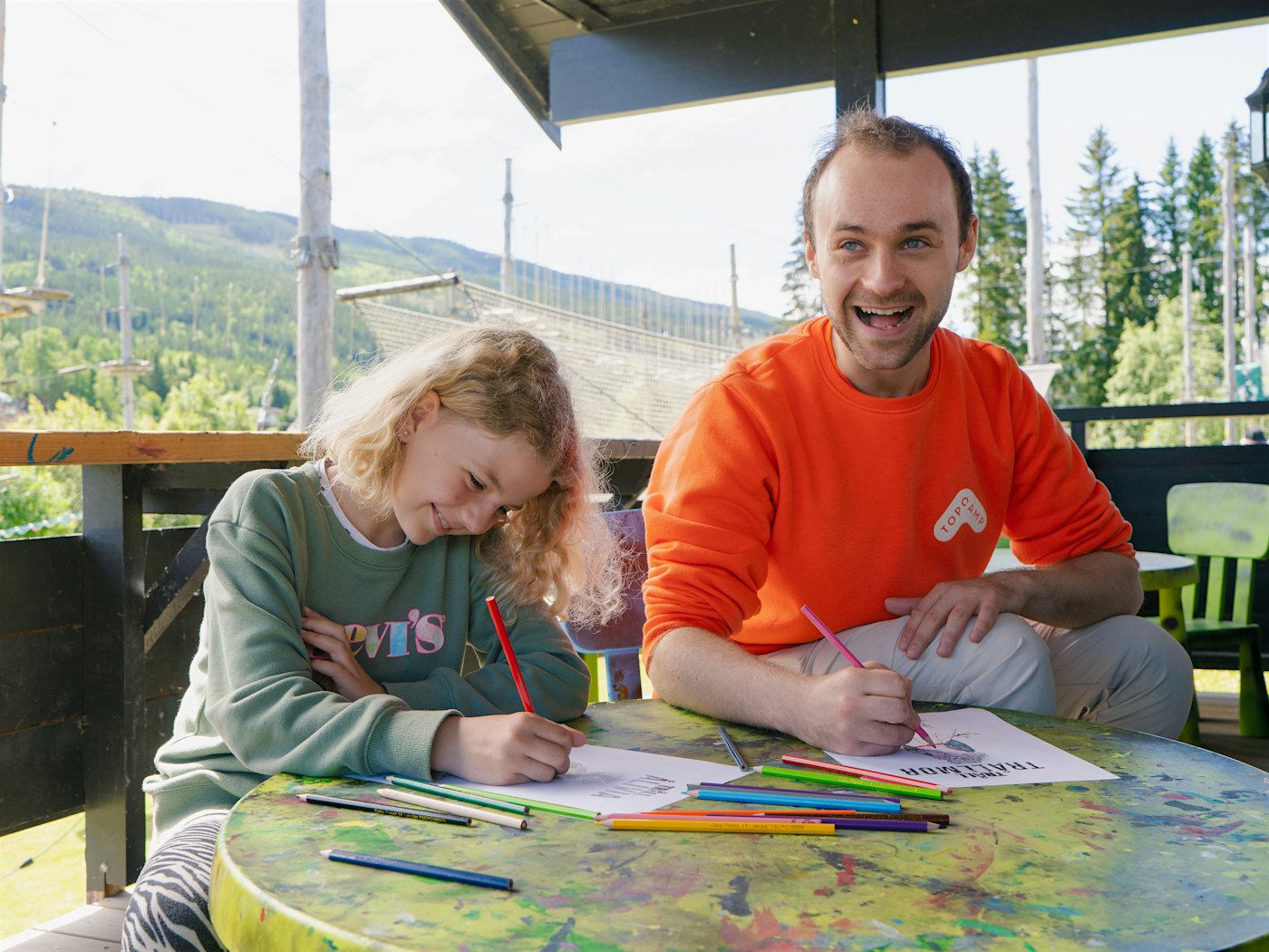 En jente og en aktivitetsleder ler og fargelegger troll-tegninger sammen. Foto