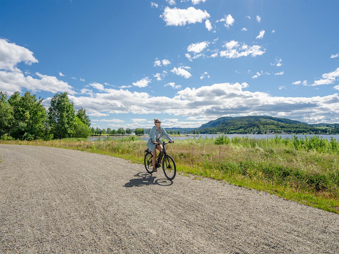 A woman cycles along a dirt road with a nice view of Mjøsa, green trees and a small flower meadow. Photo