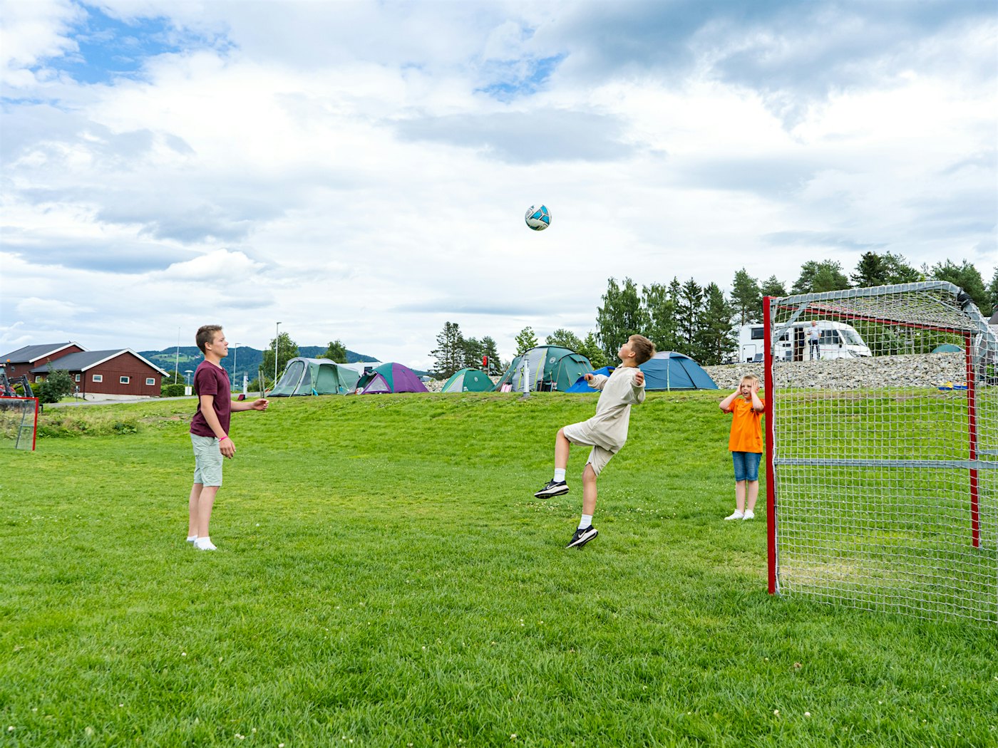 Three boys play football on a football pitch, tent in the background. Photo