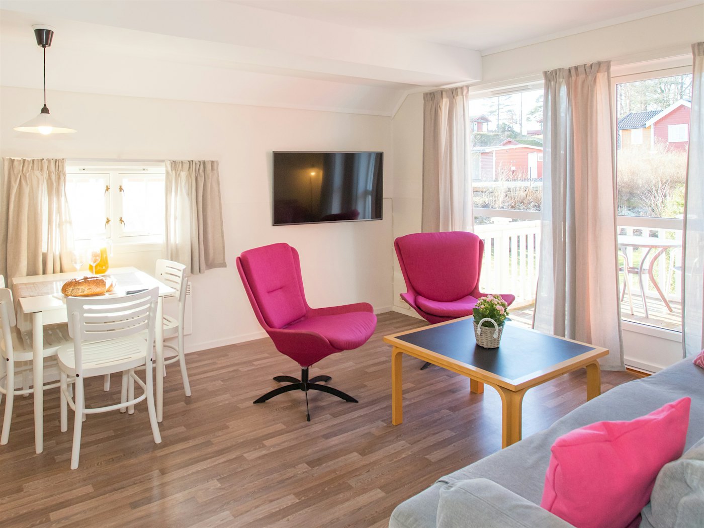 Heller und offener Raum mit viel natürlichem Licht durch große Fenster. Zwei rosafarbene Sessel, Sofa, Fernseher und gedeckter Esstisch zum Frühstück. Foto