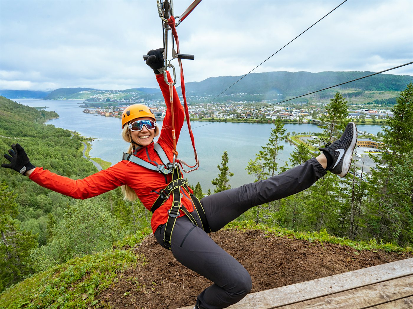 Woman hangs in a zipline with her arms out and smiles big, with a view of Mosjøen in the background. Photo