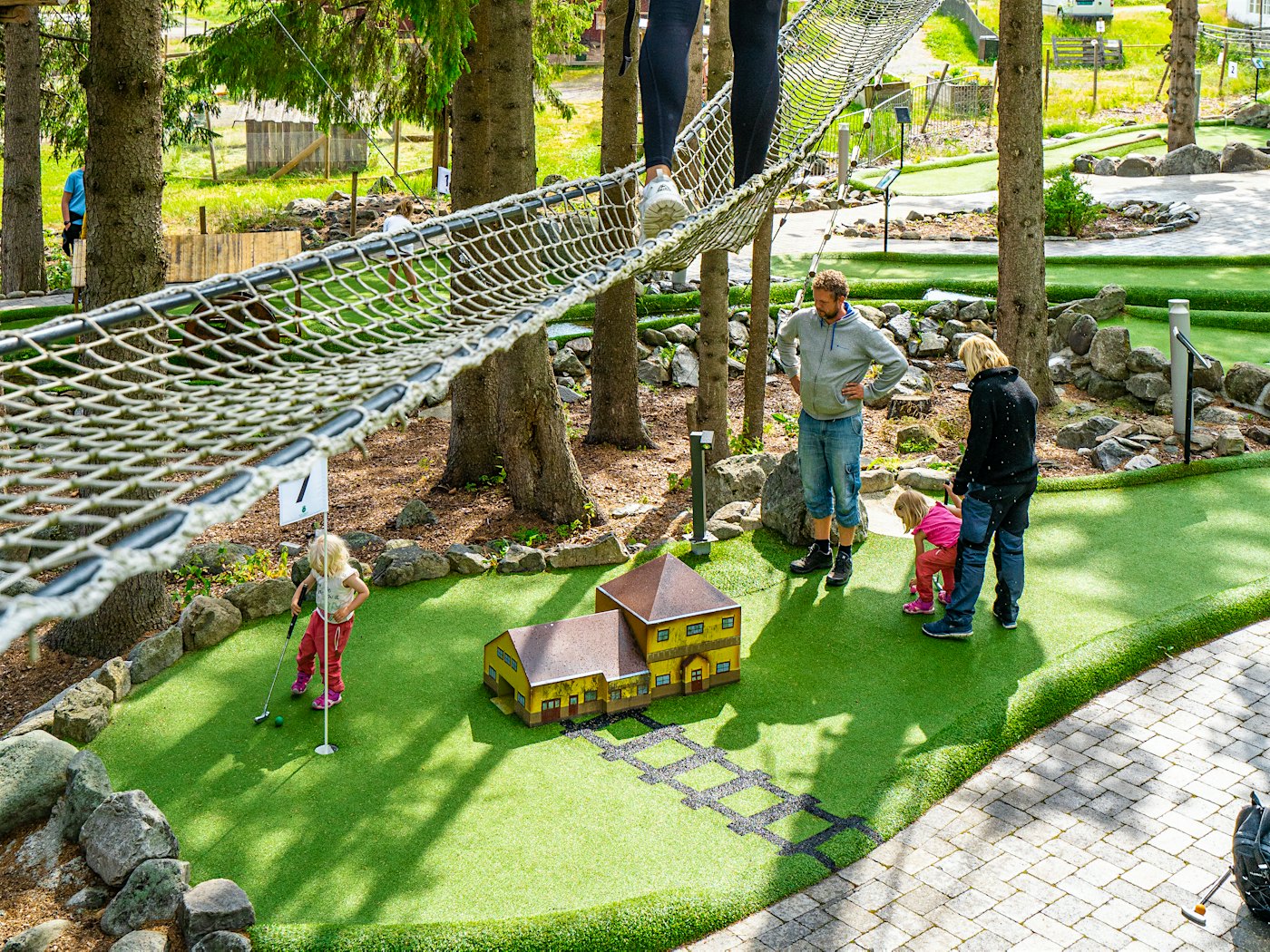 Kvinne går over netting i klatrepark mens hun ser ned på familie som spiller eventyr-minigolf på bakken. Foto
