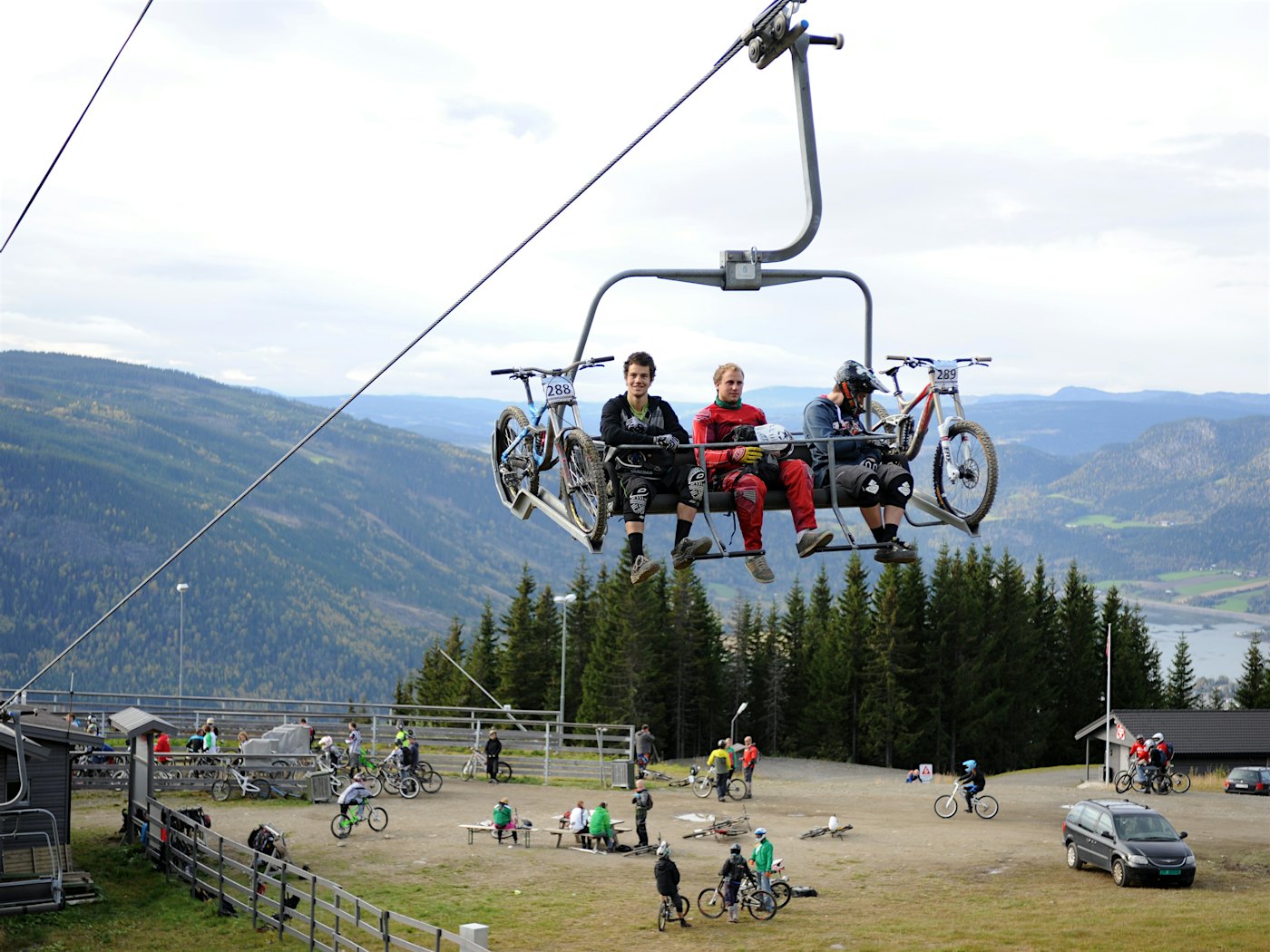 Three cyclists sit in a chairlift with their bikes, several cyclists on the ground below, mountains in the background. Photo