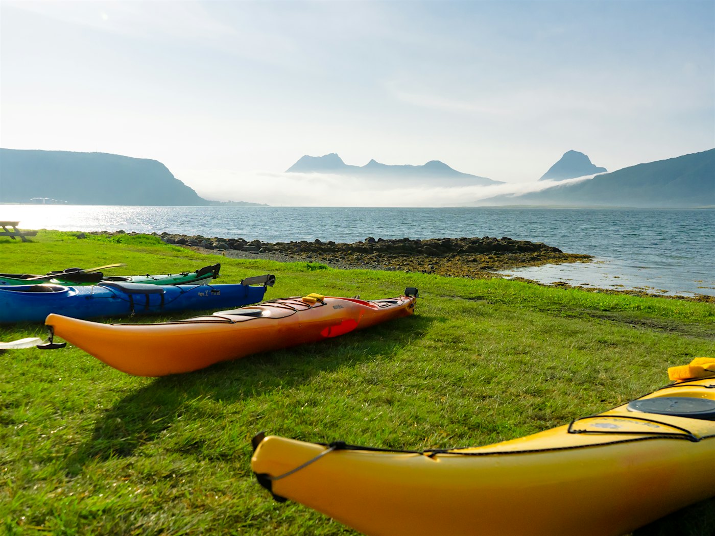 Four kayaks lie on a lawn in front of the sea, with mountains in the background. Photo