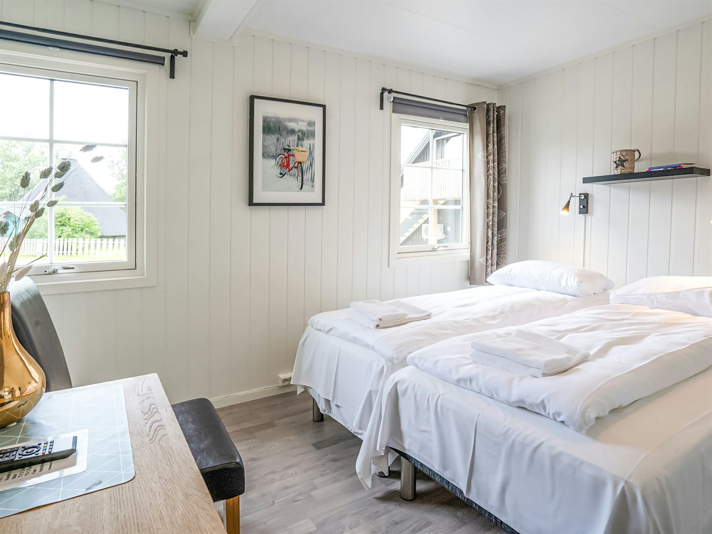 Lyst rom med dobbeltseng med hvitt sengetøy, rommet er pyntet med bilde på veggen og en vase. Foto