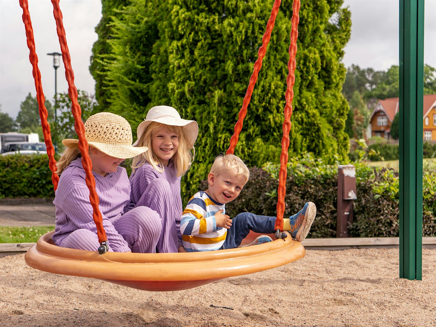 Two girls in matching outfits and their little brother sit together in a large swing on the playground. Photo