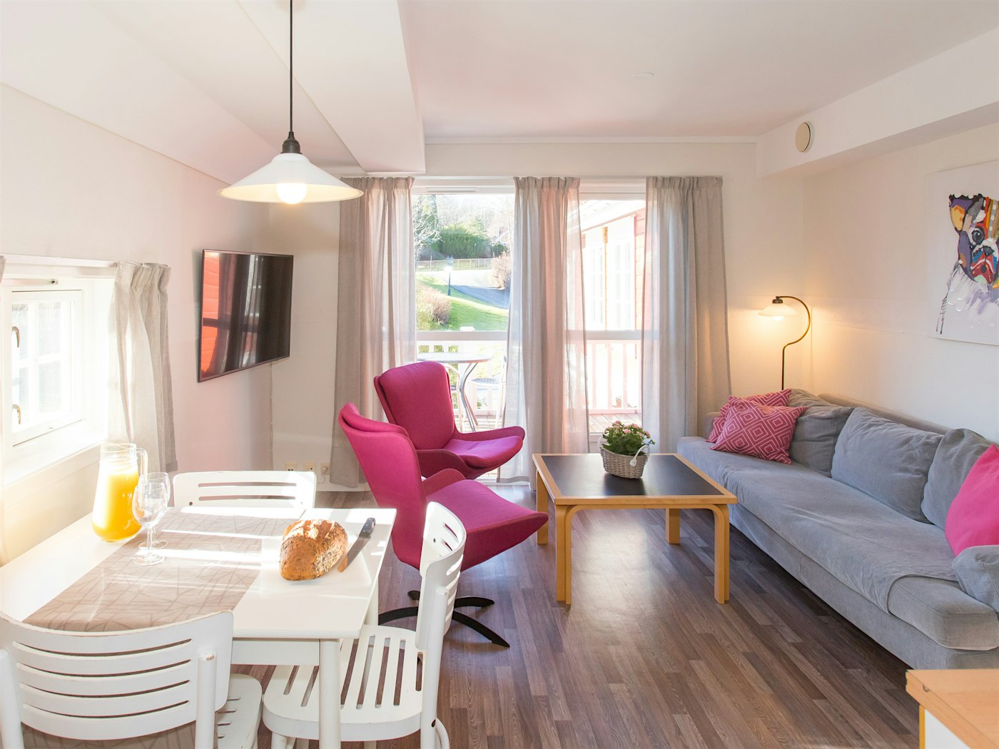 Lyst og åpent rom med mye naturlig lys fra store vinduer. To rosa lenestoler, sofa, TV og dekket spisebord for frokost. Foto