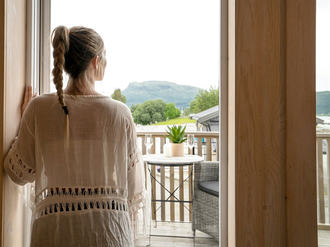 Kvinne står i vinduskarm i hytte og kikker utover fjell-utsikt. Foto