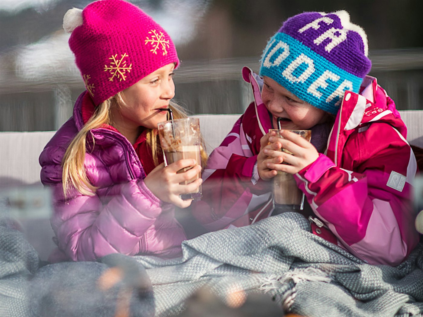Zwei Mädchen lachen und trinken Kakao am Feuer mit Decken über ihnen im Winter. Foto
