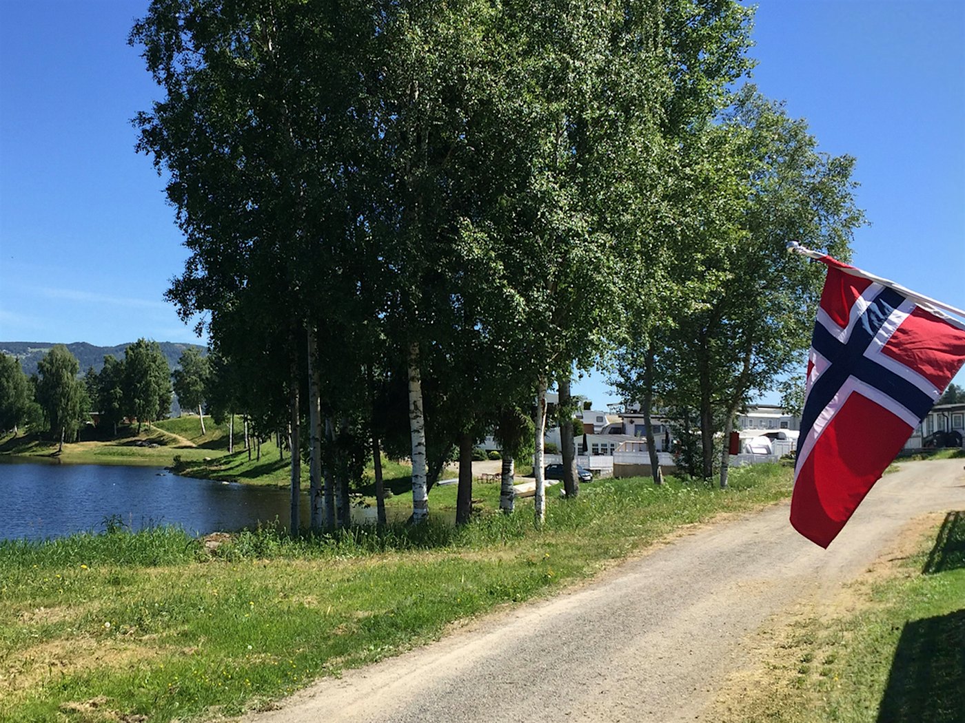 Weißer Verandazaun mit der norwegischen Flagge und Mjøsa mit seiner grünen Umgebung zu beiden Seiten eines schmalen Schotterwegs. Foto
