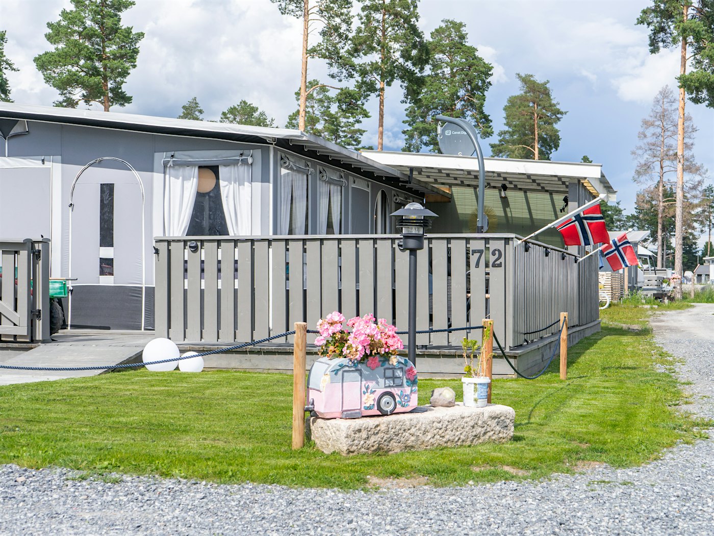 Fastplass med inngjerdet terrasse på campingplass. To Norske flagg og blomsterpotte med rosa blomster utenfor. Foto