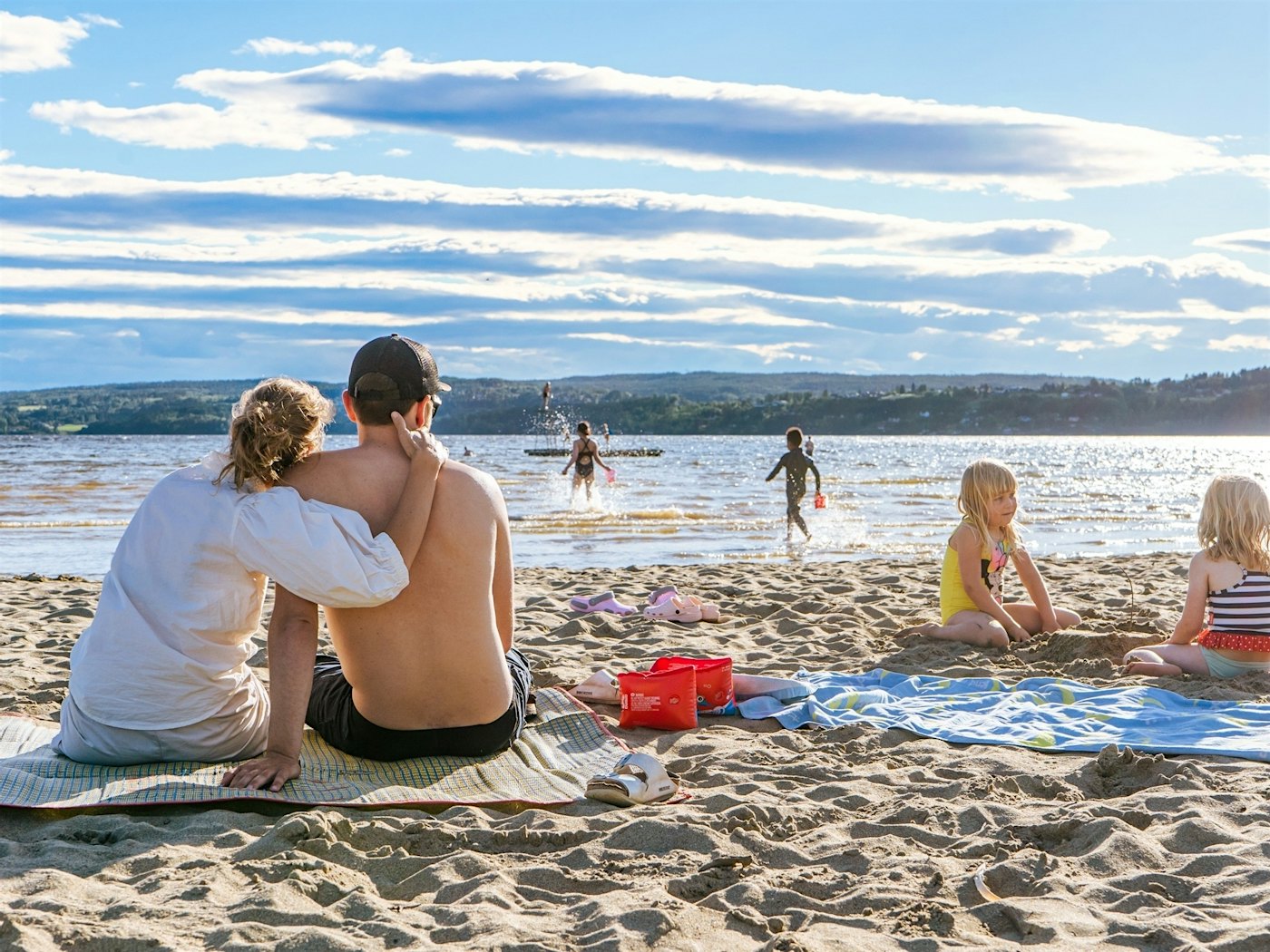 Frau und Mann sitzen auf einem Handtuch am Strand, die Frau hält den Mann um sich, während sie auf den Strand und das Wasser blicken. Zwei Mädchen spielen nebenan im Sand. Andere Kinder laufen ins Wasser. Foto